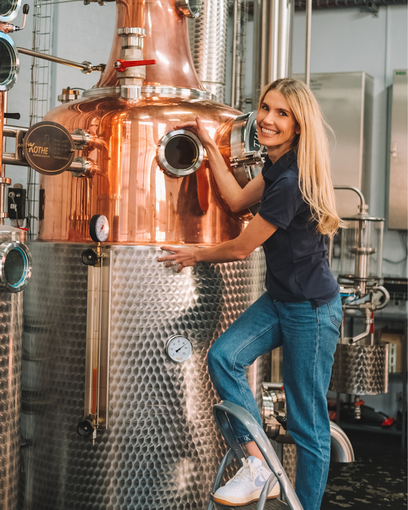 Shanna-Rae Master Distiller of Mirari Gin posing with a large copper still in a distillery setting