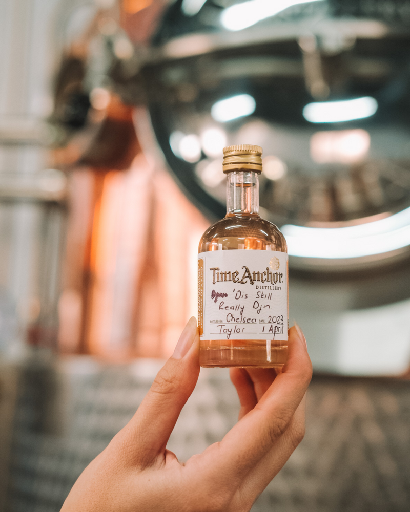 Hand holding a small bottle of Time Anchor Distillery bottle your own Gin infron t of a large copper pit still